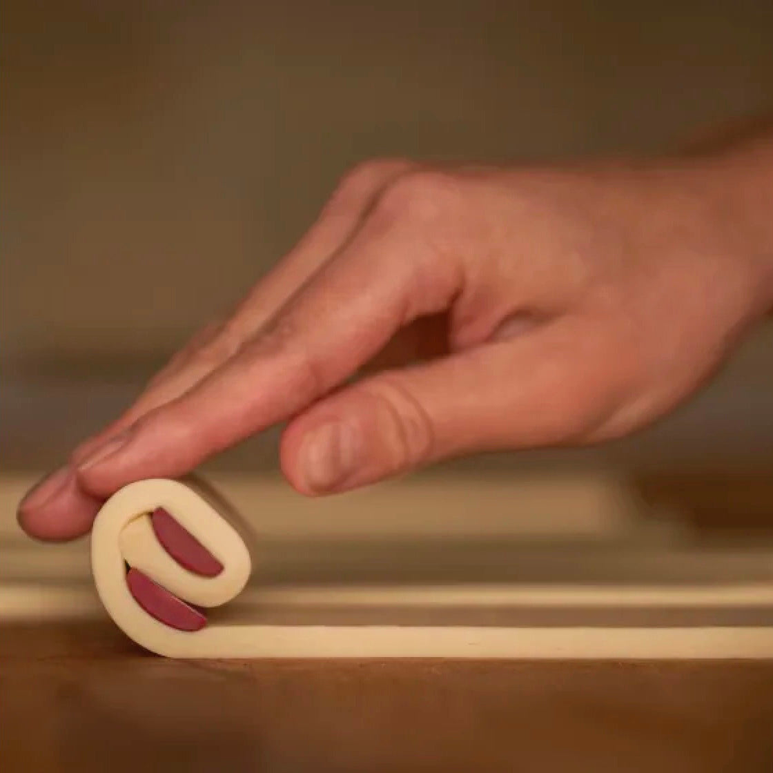 A close-up of a hand rolling out a long strip of pale dough with filling of Valrhona Raspberry Inspiration Chocolate Batons over a wooden surface, with a small rolled piece resting on top.