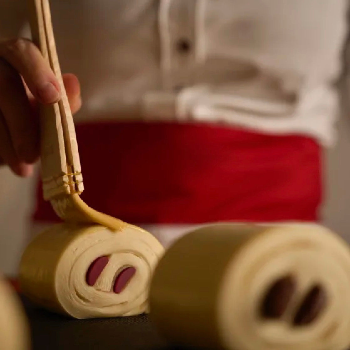 A close-up of a person wearing a white shirt and red apron using wooden chopsticks to spread a yellow sauce onto rolled dough with Valrhona Raspberry Inspiration Chocolate Batons, set against a blurred background.