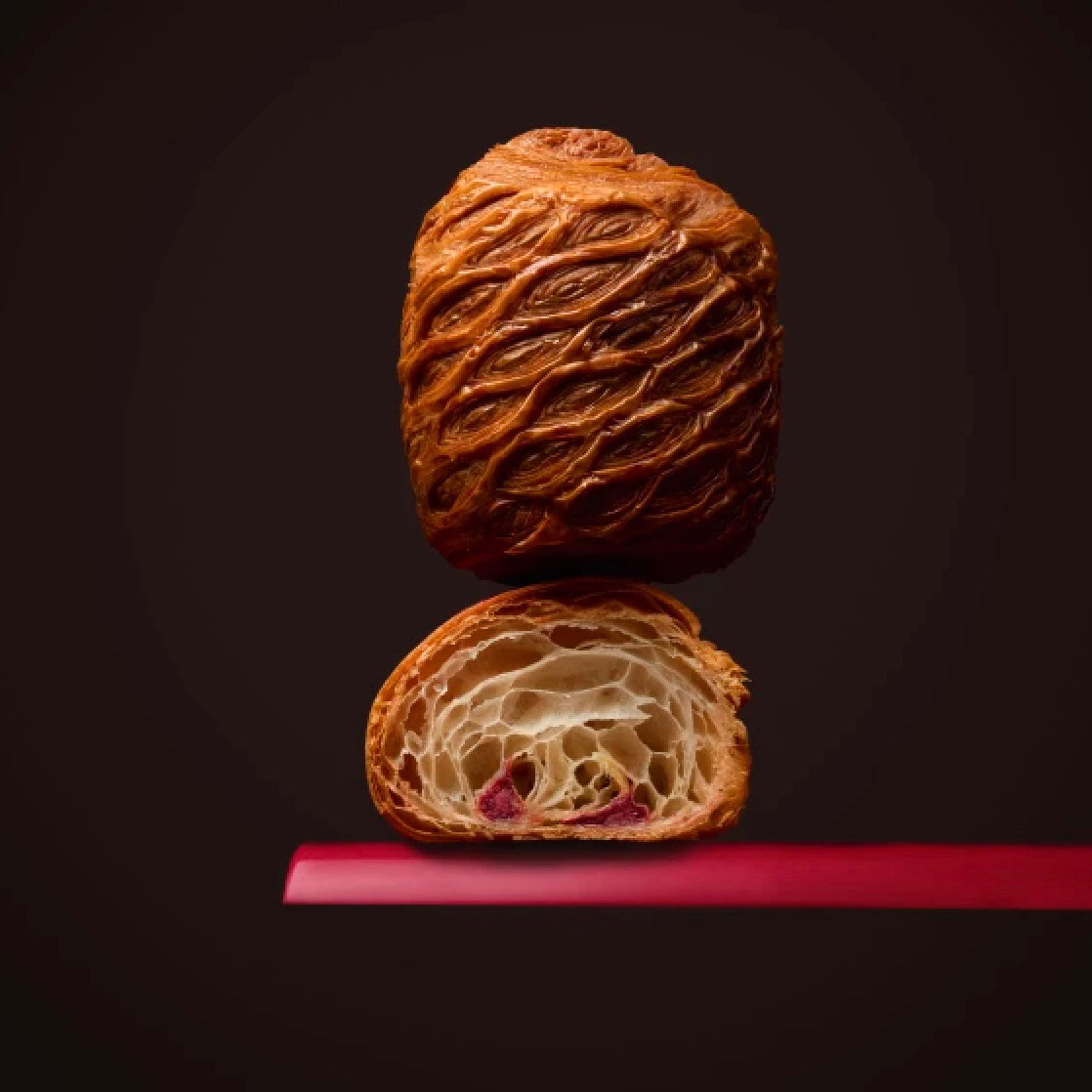 A baked pastry shown whole and sliced open on a dark studio background, with a glossy brown exterior and a light crumb interior with Valrhona Raspberry Inspiration Chocolate Batons.