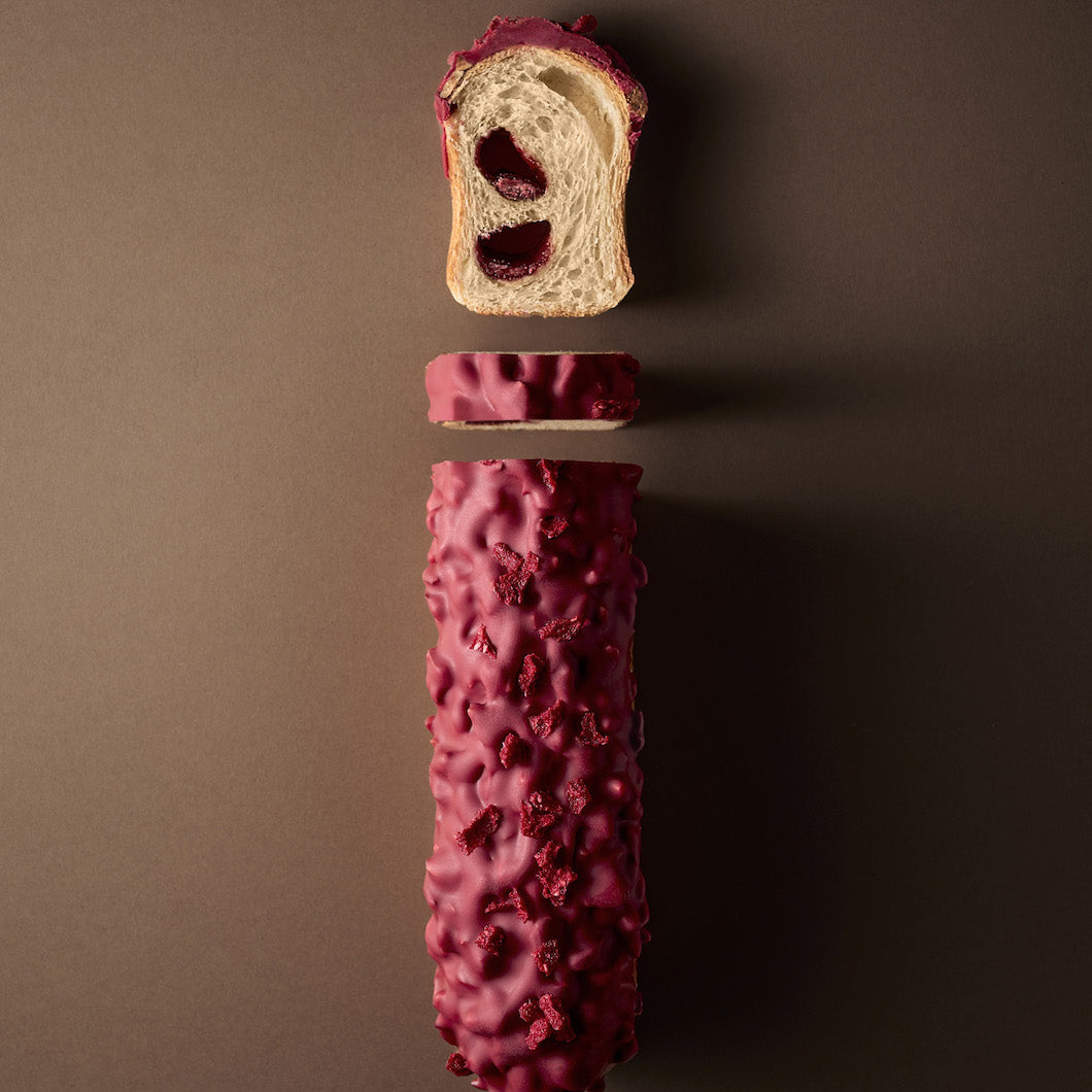 A sliced loaf of bread with a deep red berry glaze and filling of Valrhona Raspberry Inspiration Chocolate Batons, shown floating vertically against a dark brown background.