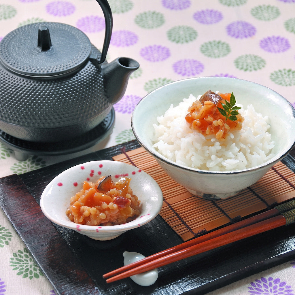 A Japanese-style meal setup with a black textured teapot, a bowl of white rice topped with a small orange-red sauce dish, a smaller side dish of the same sauce, and red-brown chopsticks on a black tray over a patterned tablecloth.
