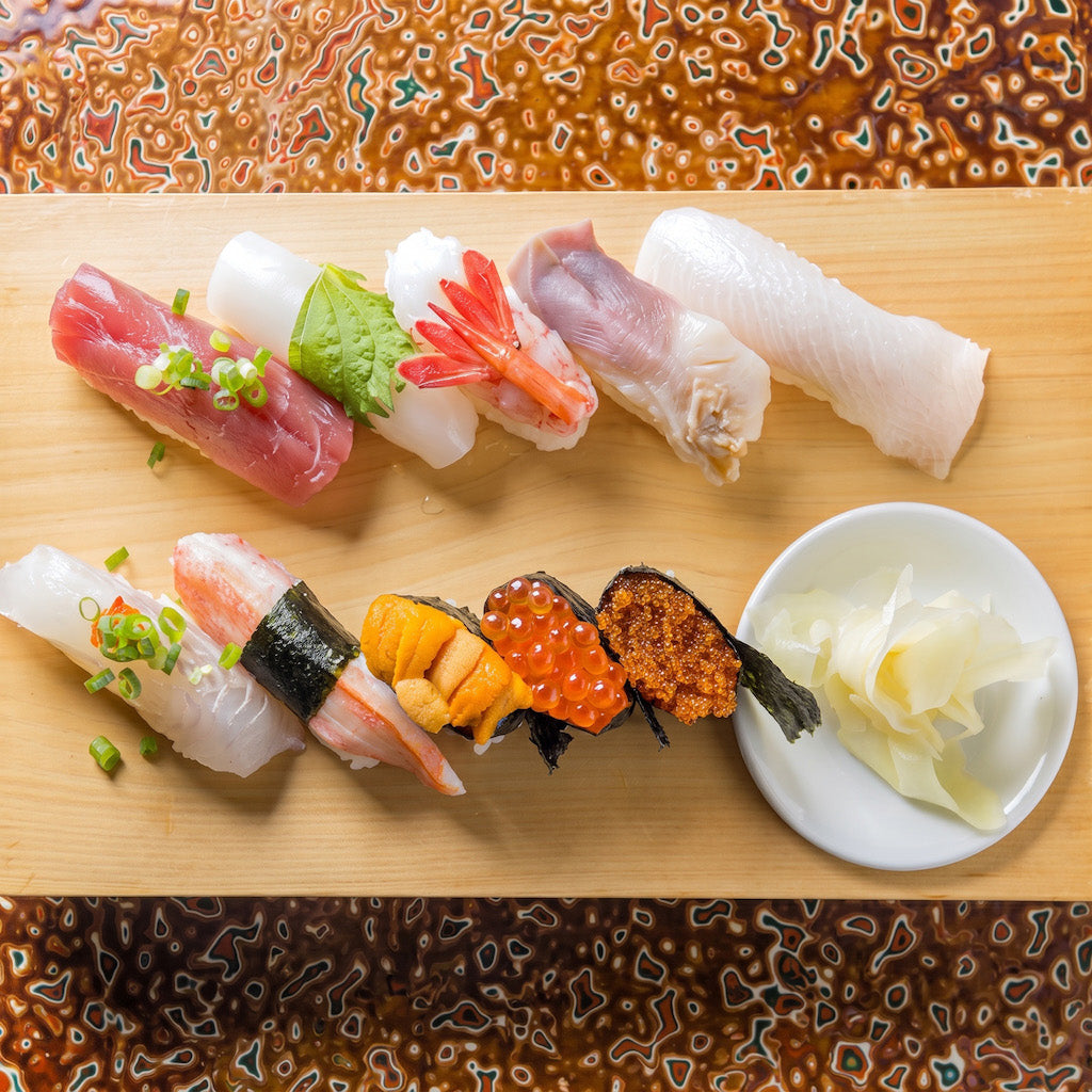 A top-down view of assorted sushi pieces arranged on a rectangular wooden board, with a small white dish of pickled ginger on the right. The sushi includes tuna, shrimp, white fish, salmon, mackerel, and other varieties, garnished with green onions and sesame seeds.