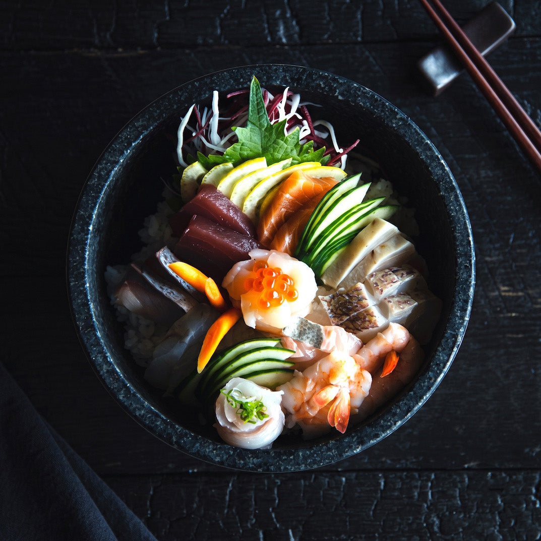 A top-down view of a black stone bowl filled with a colorful Japanese sashimi rice bowl, featuring raw fish, shrimp, cucumber, avocado, and a soft-boiled egg, set on a dark wooden table with chopsticks.