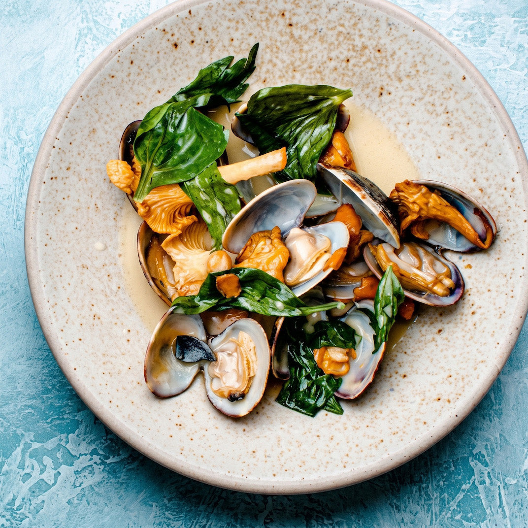 A top-down view of a seafood medallion dish, featuring mussels, clams, mussels, and mushrooms cooked with green herbs in a light sauce, served on a speckled beige ceramic plate against a textured light blue background.