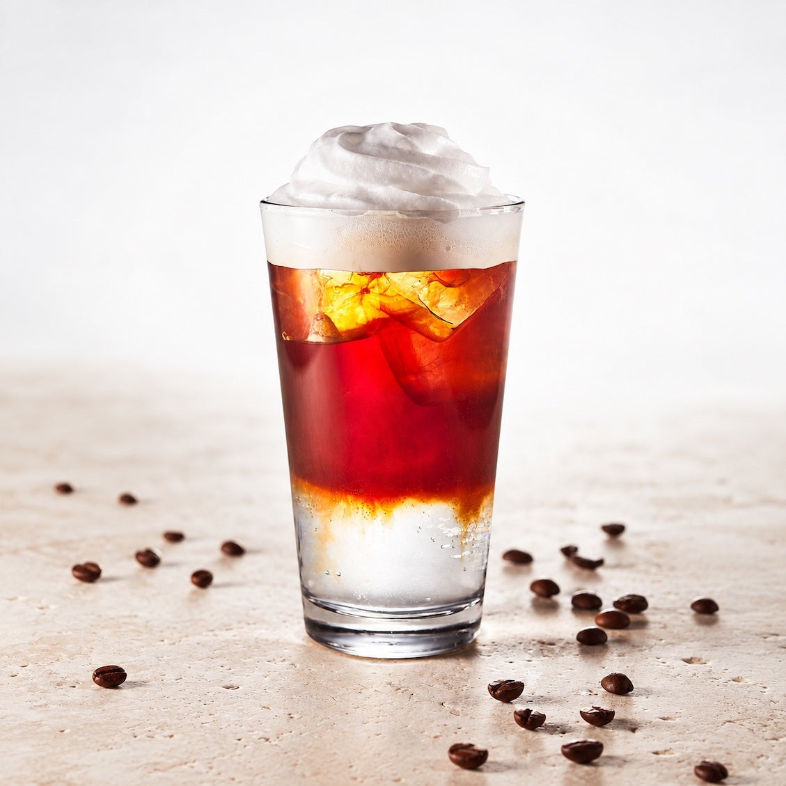 A layered iced coffee drink in a clear glass, topped with whipped cream and ice, with coffee beans scattered on a light beige surface against a white background.