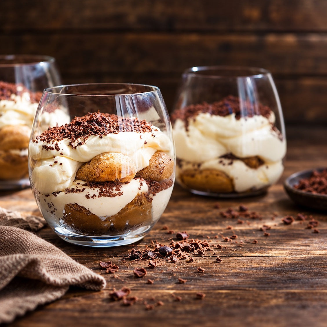 Three clear stemless wine glasses filled with layered tiramisu dessert, including cream, ladyfinger cookies, and cocoa powder sprinkles, arranged on a rustic dark wood table.