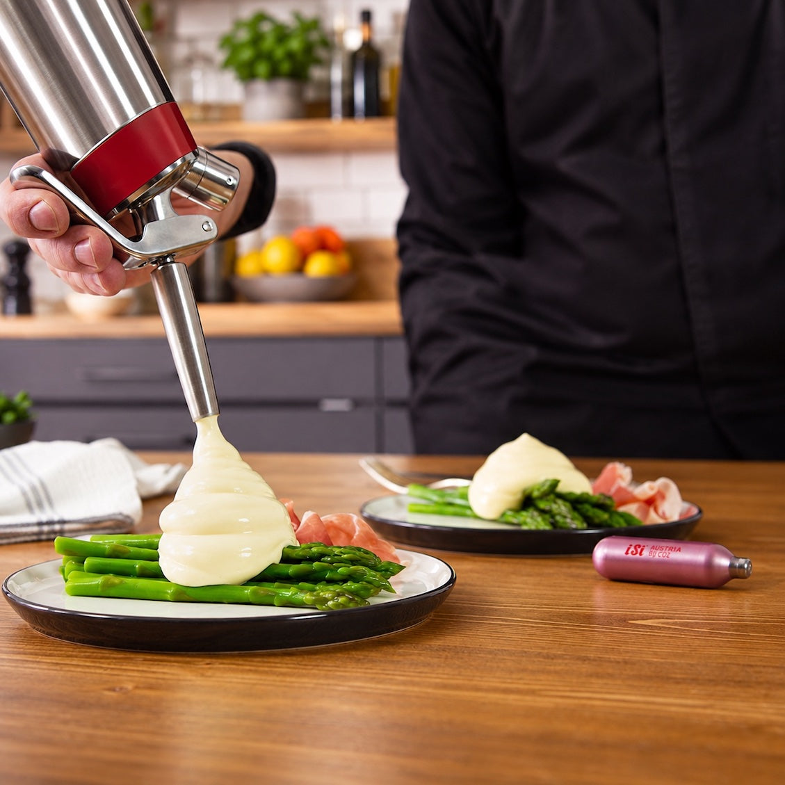 A person in a black chef coat uses a stainless steel and red squeeze dispenser to pour thick white sauce over asparagus and ham on a plate, with a matching dispenser lying on a wooden kitchen table.