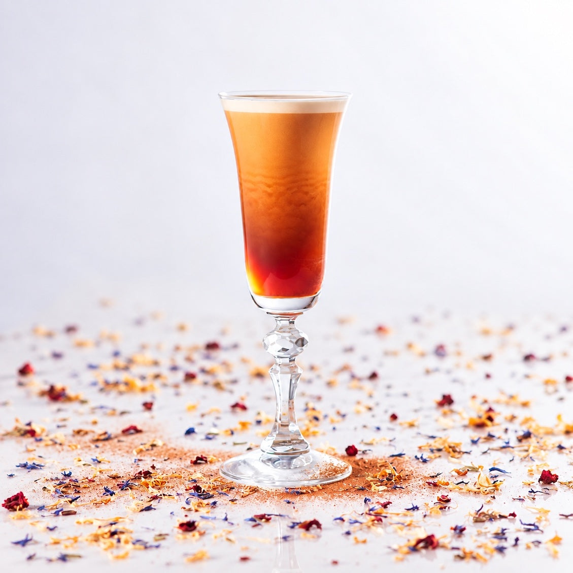 A tall, slender stemmed glass filled with an amber-orange layered drink, set on a light surface scattered with dried flowers and spices against a soft white background.